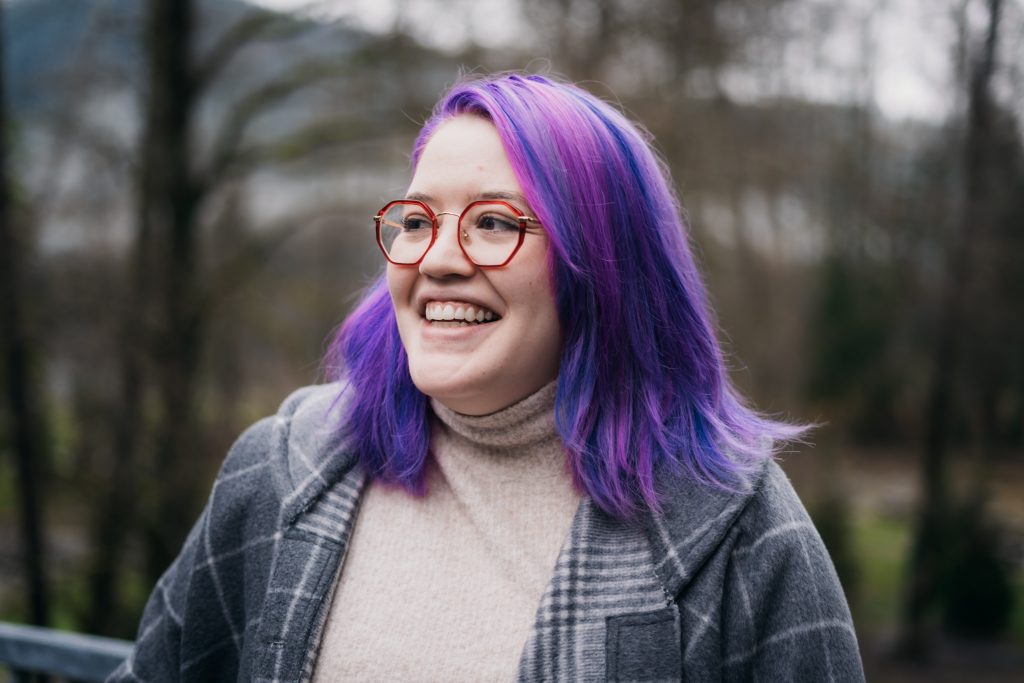 photo of Caitlyn from the bust up. She is looking off to the left and is smiling. The background is an out of focus tree and forest landscape