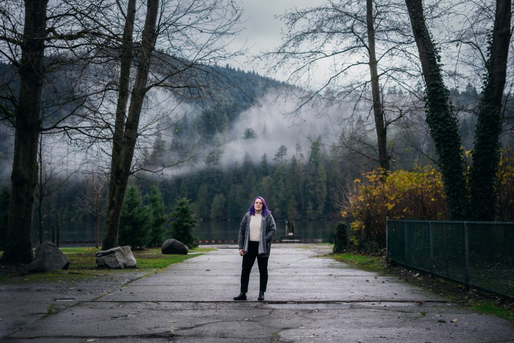 a wide-angle photo of Caitlyn amongst the trees and mountains. She stands centred on a paved path at Barnet Marine Park