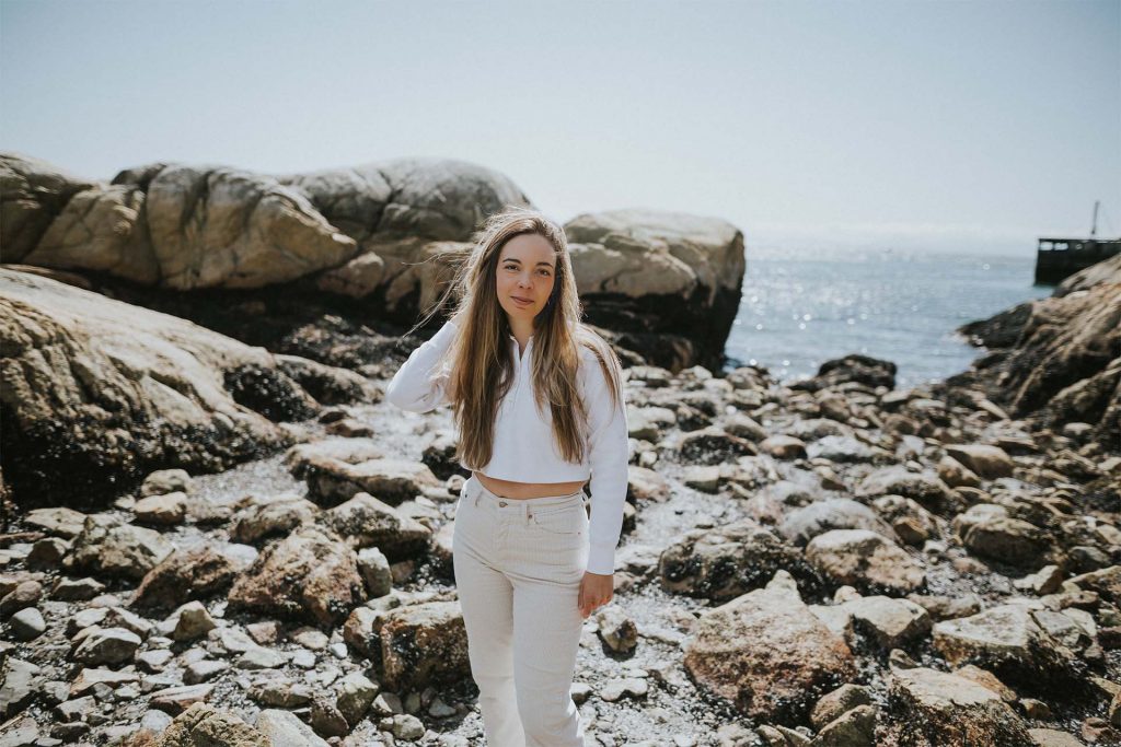 A photo of Jamie-Leigh standing on a beach with large rocks. She is looking at the camera with a slight smile