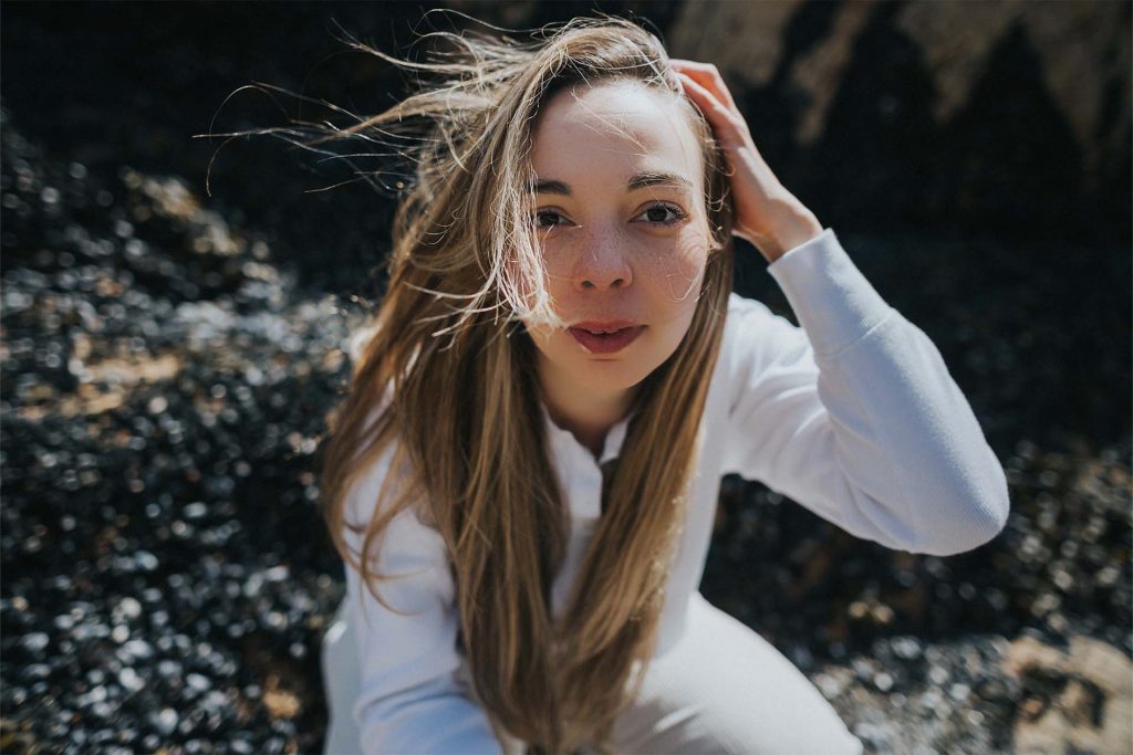 A photo of Jamie-Leigh seen kneeling on a beach with out of focus rocks and sand in the background. She is looking directly to the camera and pulling her hair back on one side. The wind is blowing her hair around her face