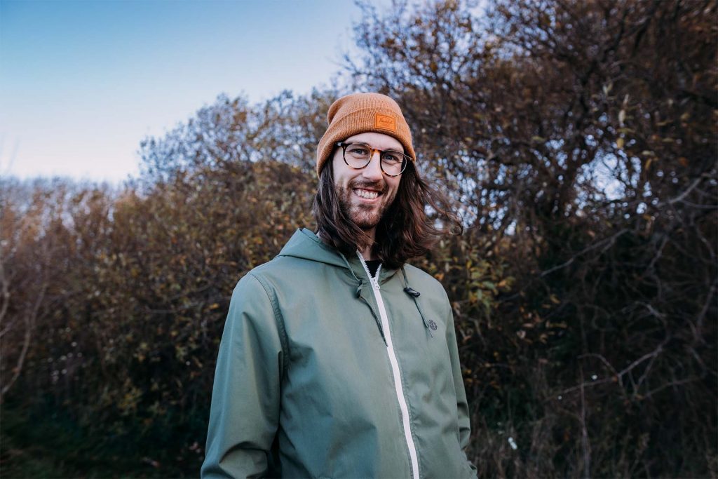 A photo of russ smiling and looking at the camera. He is surrounded by autumn trees that have shed their leaves.