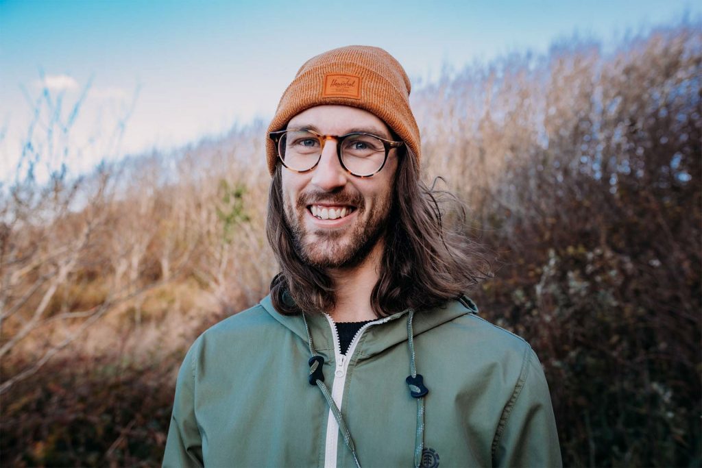 a photo of russ from the shoulders up looking at the camera smiling. He stands in front of brush and trees