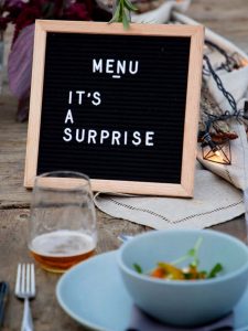 a sign reading “Menu: it’s a surprise” on a table amongst bowls of food, glasses and cutlery.
