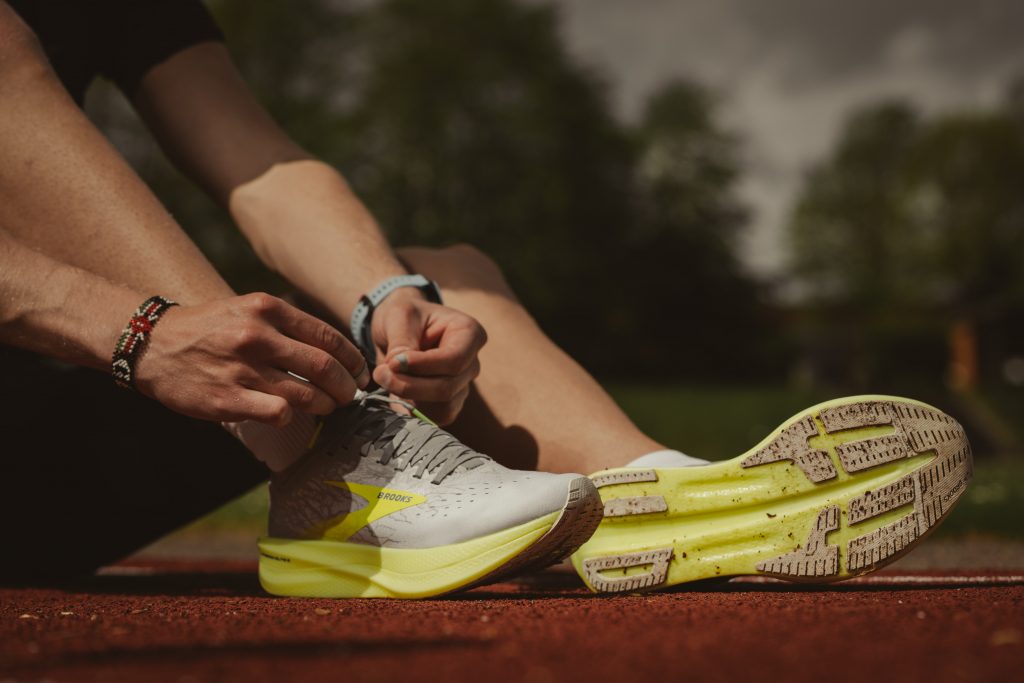 A close up of a person's legs, feet and hands. They are seated on the ground outside and are tying up their shoelaces.