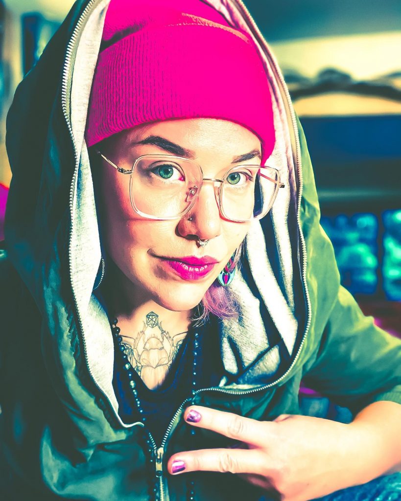 a close up of Ms.PAN!K, looking directly at the camera holding two fingers up. They are wearing a bright pink toque and matching lipstick. 