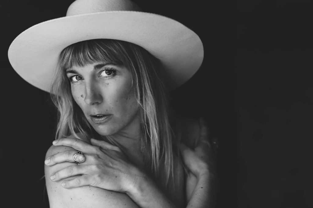 Close up of Renée Layla looking directly at the camera, hugging herself with her hands on her shoulders. She is wearing a wide brimmed hat