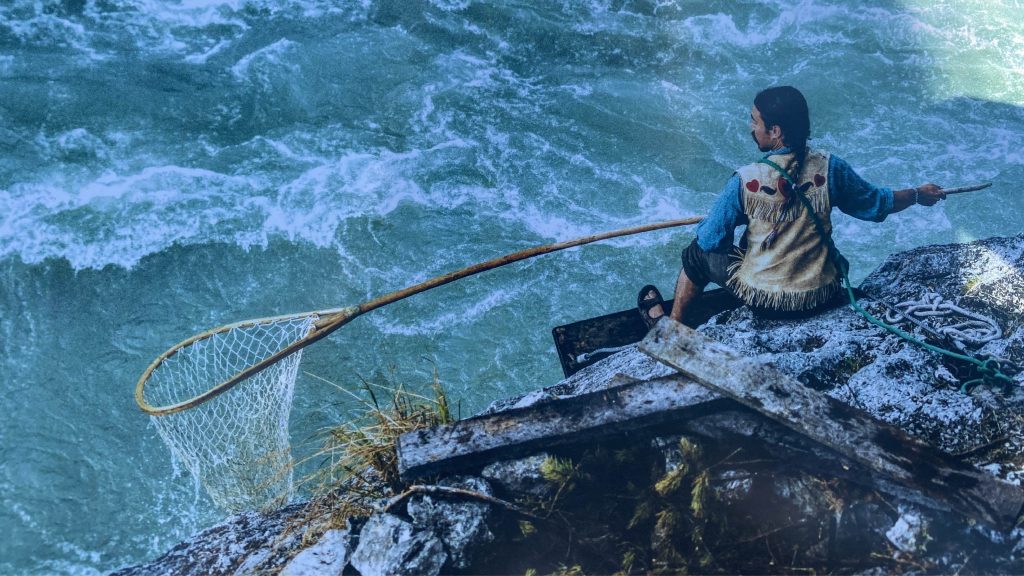 A person holding a large net sits at the edge of a rushing body of water
