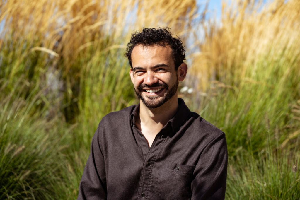 Gilberto smiling, sits in front of long grass